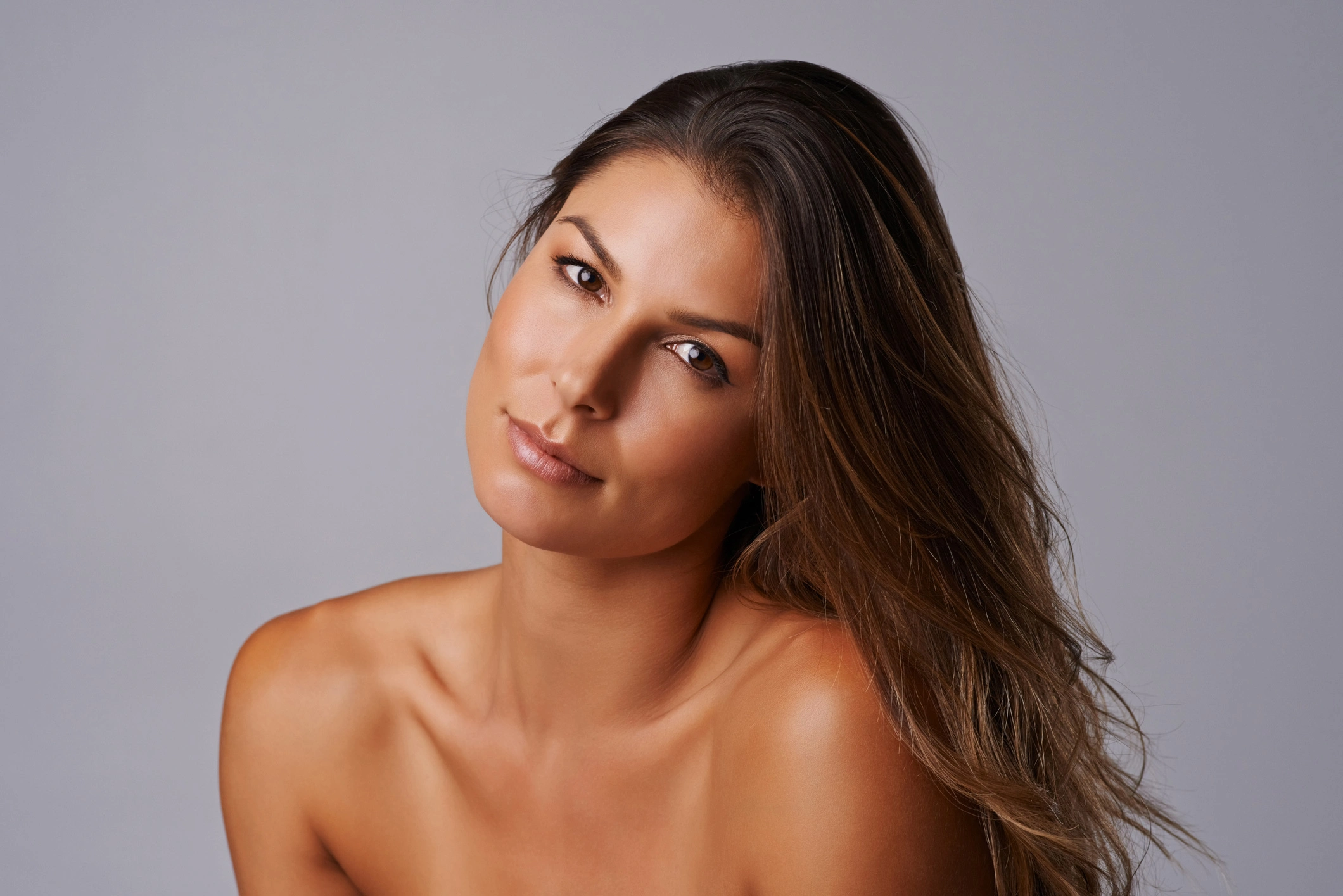 Portrait of a woman with long brown hair and sun-kissed skin, neutral gray background, beauty headshot for skincare.